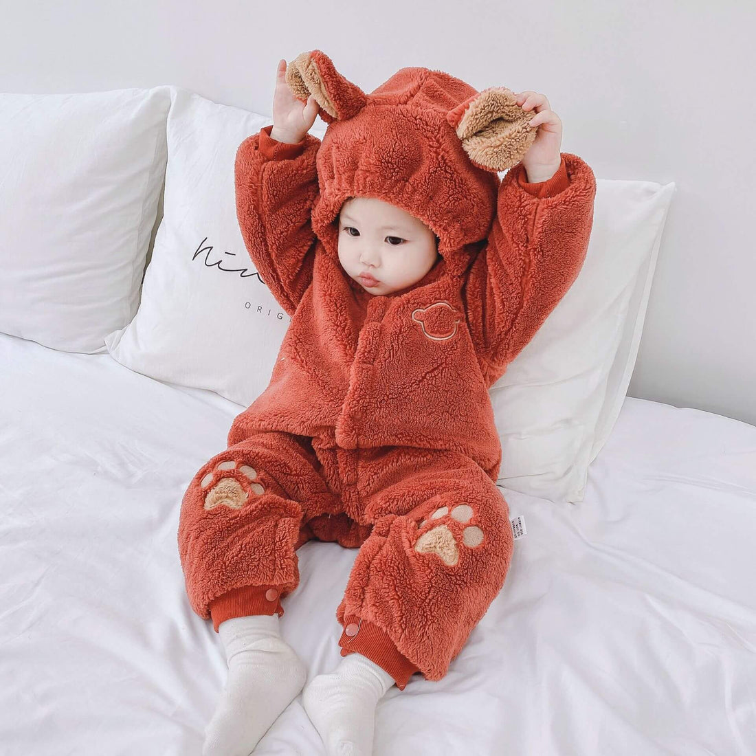 Baby in cozy orange bear costume with cute paw prints, sitting on white bed.